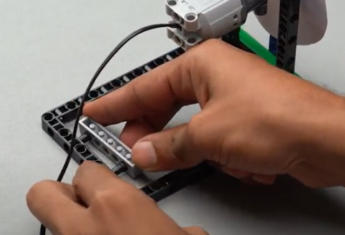 Take a 1 by 6 technic brick with holes, insert two pegs into it, and then attach it to the 15 by 7 block, to enhance the structure of our spin wheel robot, a beginner-friendly STEM robotics project.