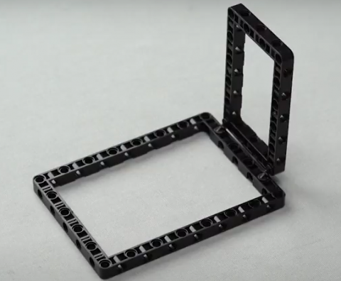 Upon the 15 by 11 technic block placed up against a grey background another technic block of 11 by 7 is attached. Two 90 degree technic pegs are used for attaching. this builds the frame for crafting our spin wheel robot, robotic project.